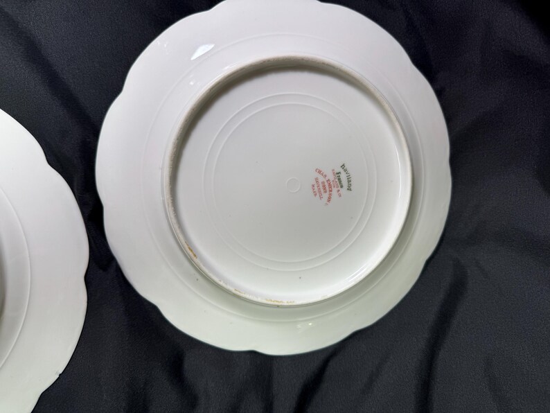 May include: Two white ceramic plates with scalloped edges. The plates have a circular design in the center and a red stamp with text. The plates are on a black surface.