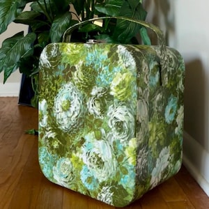 Pode incluir: Uma mala de viagem rígida de formato quadrado com um padrão floral vibrante em tons de verde, branco e azul claro. A mala tem cantos arredondados, uma pega de transporte superior e um fecho de metal prateado na borda superior.