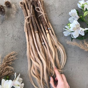 Può includere: Un set di dreadlock sintetici caratterizzati da una sfumatura di colore che va dal marrone scuro alle radici al biondo chiaro sulle punte. I dreadlock sono legati con spago sottile e decorati con piccole perline metalliche e fili avvolti.
