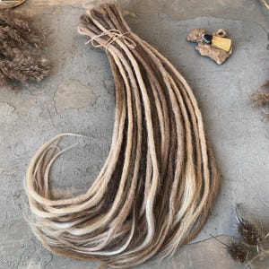 Peut inclure: Un faisceau de dreadlocks synthétiques présentant une palette de couleurs dégradées, passant du brun foncé aux racines au blond clair aux pointes. Les mèches sont attachées ensemble au sommet par un cordon fin, formant une longue extension capillaire.