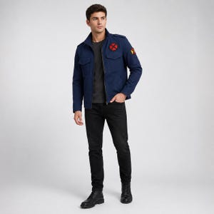 Könnte beinhalten: Eine dunkelblaue Utility-Jacke mit Stehkragen, Reißverschluss, zwei Brusttaschen und zwei runden Aufnähern mit roten und gelben Motiven auf Brust und Ärmel. Die Jacke wird über einem dunkelgrauen T-Shirt und einer schwarzen Jeans getragen.
