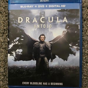 Può includere: Una custodia blu per Blu-ray del film Dracula Untold. La copertina presenta una figura centrale in armatura scura con un grande mantello, un castello sullo sfondo e il testo Dracula Untold e Every Bloodline Has A Beginning.