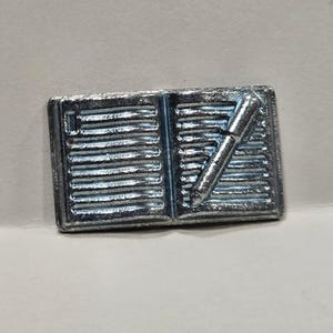 Può includere: Una spilla in metallo color argento a forma di libro aperto con pagine a righe. Una penna è raffigurata in diagonale sulla pagina destra del libro. La superficie della spilla ha una lucentezza metallica con dettagli in rilievo definiti.