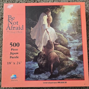 Puede incluir: Una caja de rompecabezas de 500 piezas titulada Be Not Afraid de Greg Olsen, que mide 45,72 cm por 60,96 cm. La caja presenta una pintura de una figura con túnica blanca ayudando a un niño a trepar rocas cerca de un arroyo, con el logotipo de SunSout.