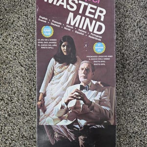 May include: A rectangular game box for Super Master Mind by Invicta. The dark brown box features a photograph of a person in a white sari and a person in a white suit with a bowtie. Text includes "Super Master Mind," "A game of cunning and logic for two players," and "Now five holes and eight colours. Up to 59,000 permutations."