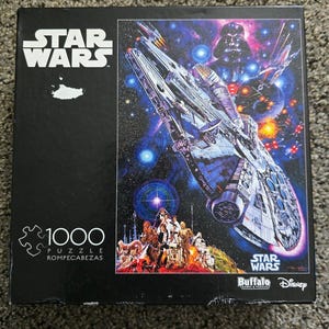 Może przedstawiać: Kwadratowe pudełko puzzli Star Wars składające się z 1000 elementów, przedstawiające scenę kosmiczną z Sokołem Millennium, Darthem Vaderem i różnymi postaciami. Czarne pudełko zawiera logo Star Wars, liczbę elementów oraz marki Buffalo Games i Disney.