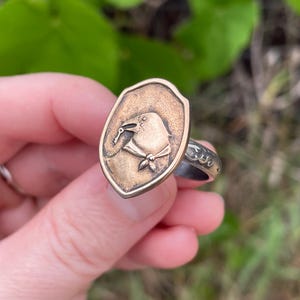 May include: A metal ring featuring a shield-shaped face with a raised relief design of a bird wearing a neckerchief. The band of the ring has a textured, patterned surface. The metal has a warm, aged bronze tone with darker recessed details.