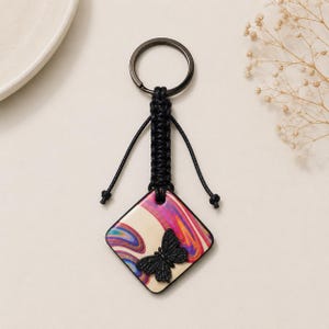 Puede incluir: Un llavero con un colgante cuadrado con un patrón de remolinos jaspeados en crema, rosa y morado. Una pequeña mariposa negra tridimensional está fijada en la parte frontal. El colgante cuelga de un cordón de macramé negro y una anilla de metal oscuro.
