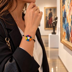 Puede incluir: Una pulsera con un dije circular con un patrón de vitral multicolor en azul, amarillo, naranja y rojo, centrado con un segmento blanco. El dije está unido a un cordón negro fino con pequeños detalles de metal en tono dorado a cada lado.