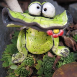Könnte beinhalten: Eine Keramikfroschfigur mit einem leuchtend lindgrünen Körper, einem breiten Maul und großen, hervorstehenden weißen Augen. Der Frosch hat dunkelgraue Akzente an den Gliedmaßen und am Maul und hält eine kleine rote und gelbe Blume im Mund.