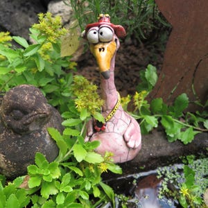 Könnte beinhalten: Eine Keramik-Gartenfigur in Form eines rosa Vogels mit rissiger Glasur, der einen roten Hut und eine gelbe Halskette mit schwarzen Punkten trägt, neben einer dunklen, abgerundeten und strukturierten vogelähnlichen Figur im grünen Blattwerk.