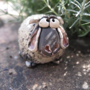 Könnte beinhalten: Eine kleine, runde Schaf-Figur aus Keramik mit einem strukturierten, cremeweißen Körper und dunkelgrauen Sprenkeln. Das Schaf hat ein glattes, dunkelgraues Gesicht, große hervorstehende Augen und zwei Schlappohren mit rosa Details.