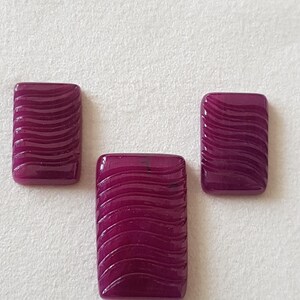 May include: Three rectangular magenta-colored cabochons with rounded corners and a carved wavy, horizontal line pattern across the surface. The pieces are arranged with one larger central piece and two smaller pieces on either side.