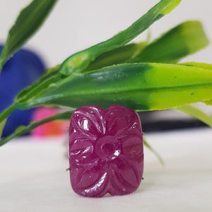 May include: A rectangular, deep magenta-colored gemstone featuring a carved floral pattern on its surface. The stone has a smooth, polished finish with distinct, recessed lines forming the petals of a flower design centered on the face.