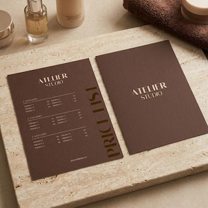 May include: Two dark brown rectangular cards featuring the text ATELIER STUDIO in gold lettering. One card displays a price list with categories and product names, while the other serves as a cover. The cards are placed on a textured stone surface.