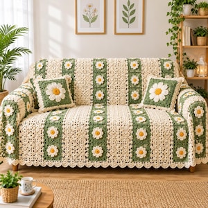 Puede incluir: Una funda de sofá de ganchillo y dos cojines cuadrados a juego con un patrón repetitivo de motivos de margaritas color crema con centros amarillos, intercalados con franjas verticales de cuadrados de ganchillo verde oscuro y borde festoneado.