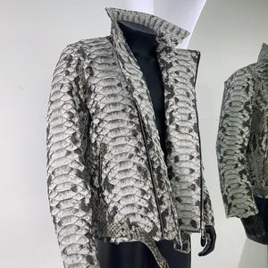 May include: A jacket featuring a textured snakeskin pattern in shades of grey, white, and black. The garment includes a high collar, a full-length front zip, and a matching belt at the waist with a metal buckle, displayed on a mannequin.