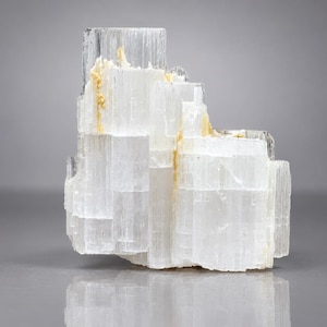 Puede incluir: Un racimo de cristales de selenita blanco translúcido con una formación escalonada y bloqueada. El espécimen mineral presenta estrías verticales e inclusiones minerales de color amarillo pálido natural en las grietas de la estructura.