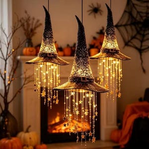 Op de afbeelding: Drie zwarte heksenhoedlampen die aan het plafond hangen. Elke hoed heeft een textuur, een gespdetail en warm gloeiende LED-lichtsnoeren die naar beneden vallen, versierd met kleine stervormige ornamenten en doorzichtige kralen.