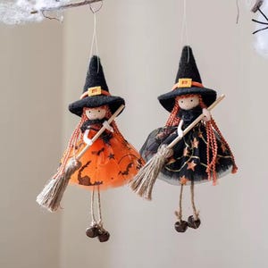 Può includere: Due bambole streghe appese con abiti in tulle arancione e nero, cappelli a punta neri con fasce arancioni e lunghi capelli in filato arancione. Ogni bambola tiene una piccola scopa in legno chiaro e fibre naturali, con campanellini ai piedi.