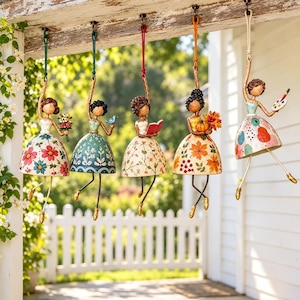 May include: Five hanging bell ornaments shaped like figures with dresses. The first has a floral dress, the second a green dress with a bird, the third a dress with vines and a book, the fourth a dress with autumn leaves and a pumpkin, and the fifth a patterned dress with a paint palette.