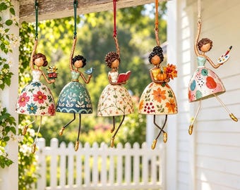 May include: Five hanging figurines shaped like dancing figures with bell-shaped skirts. Each figure features a unique painted design, including flowers, leaves, a book, a pumpkin, and an artist palette, suspended by colourful cords.