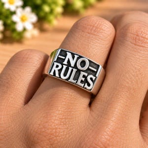 Op de afbeelding: Een zilverkleurige metalen ring met een rechthoekig voorvlak waarin de tekst NO RULES is gegraveerd in dikke, zwart ingevulde hoofdletters. De ring heeft een gladde, gepolijste afwerking en een breed, plat ontwerp op de vinger.