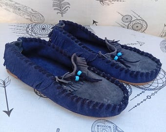 May include: A pair of dark blue suede moccasins featuring a whip-stitched edge, a fringed tongue, and decorative dark grey laces accented with two small, bright turquoise beads on the front of each shoe.