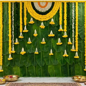 May include: A decorative backdrop featuring overlapping green banana leaves adorned with vertical garlands of yellow marigolds and white jasmine flowers. A semi-circular floral arrangement hangs at the top centre, flanked by brass oil lamps.