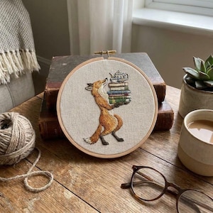May include: A circular wooden embroidery hoop containing a beige fabric panel with a hand-stitched design of an orange fox standing on its hind legs while balancing a stack of colorful books and a small teapot on its head.