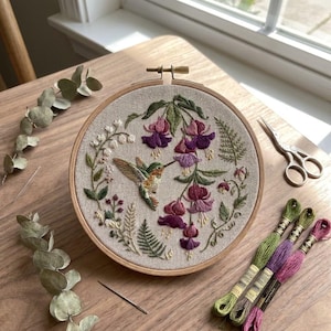 May include: A circular wooden embroidery hoop containing a beige fabric panel with a hand-stitched design of a hummingbird feeding on purple and pink fuchsia flowers, surrounded by white lily of the valley, green ferns, and small berries.