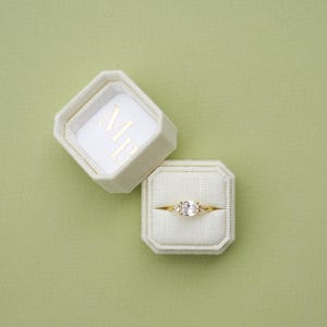 May include: A gold ring with a clear oval-cut center stone and two smaller side stones, resting in a cream-colored octagonal velvet ring box. The open lid of the box displays the gold embossed letters MB on a white fabric interior lining.
