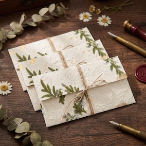 Puede incluir: Tres sobres rectangulares de papel artesanal texturizado en color blanco roto, decorados con hojas de helecho verdes prensadas y copos de pan de oro. Los sobres están atados con un cordel marrón fino en un lazo sencillo.