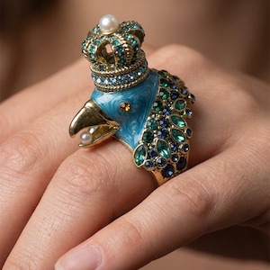 May include: A gold-tone ring featuring a bird head with light blue enamel, a crown topped with a white pearl, and a small pearl in its beak. The bird is adorned with green and blue crystals along the side, creating a detailed and ornate appearance.