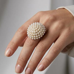 May include: A dome-shaped ring featuring a dense cluster of small, round, white pearls set closely together on a metallic base. The surface of the ring is entirely covered in these spherical white beads, creating a textured, rounded appearance.