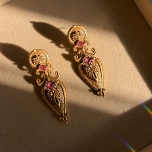 May include: A pair of gold-toned drop earrings featuring an intricate filigree design with a textured finish. Each earring is adorned with two square-cut pink stones, one set in the circular top section and one in the elongated teardrop base.