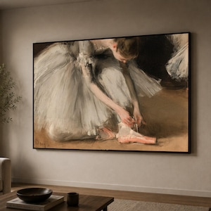 May include: A framed rectangular wall art print depicting a ballet dancer in a voluminous white tutu, leaning forward to adjust a pink ballet slipper. The scene is rendered in soft, muted tones with a dark background and a warm, earthy floor.