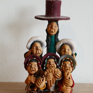 以下が含まれることがあります： 6体の人物が彫刻された手塗りの粘土製キャンドルホルダー。一番上の人物がマルーン色のキャンドルカップを支えています。人物は色鮮やかな帽子と伝統的な衣装を身にまとい、中央の人物はパンフルートを演奏し、他の人物も楽器を手にしています。