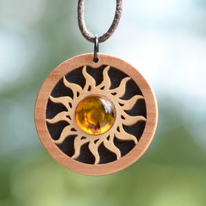 Puede incluir: Un colgante circular de madera con un diseño de sol tallado con rayos ondulados que rodean un cabochon central pulido de color ámbar. El colgante está suspendido de un cordón marrón, mostrando un fondo oscuro detrás del sol de madera clara.