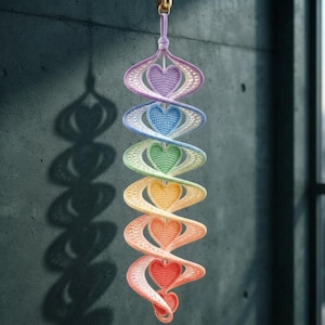 May include: A vertical hanging decoration featuring a spiral structure made of crocheted yarn in a rainbow gradient. The spiral contains six solid crocheted hearts in purple, blue, green, yellow, orange, and red, suspended from a small metal ring.