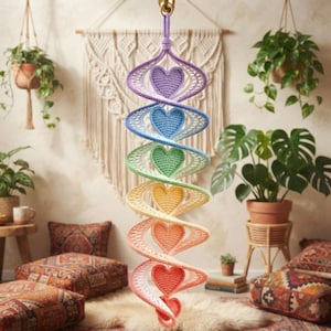 May include: A hanging spiral mobile featuring a vertical sequence of six crocheted hearts in a gradient of colors including purple, blue, green, yellow, orange, and pink. The hearts are connected by a continuous, spiraling, open-weave crochet structure.