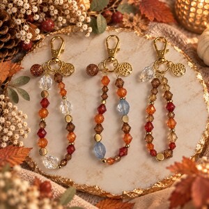 May include: Three beaded keychains featuring gold-tone lobster clasps and tree of life charms. Each keychain is strung with a variety of faceted glass and metallic beads in shades of amber, brown, red, clear, and blue, arranged in loops.
