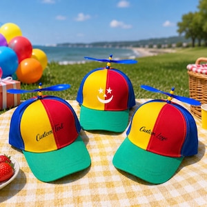 May include: Three multi-colored propeller hats featuring a yellow, red, and blue panel design with a green brim. The hats are topped with a blue plastic propeller. One hat displays a white crescent moon and stars, while two feature custom text.