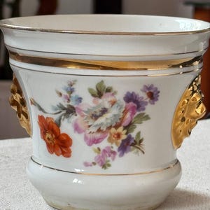 Puede incluir: Una maceta de porcelana blanca con un ramo de flores pintado a mano en tonos naranja, rosa y morado. El recipiente presenta bordes dorados en la parte superior e inferior, y cuenta con dos asas laterales en forma de cabeza de león dorada.