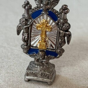 Puede incluir: Un pequeño adorno religioso de metal plateado con una cruz dorada central sobre un fondo plateado de rayos de sol. El marco está acentuado con esmalte azul, rematado con un pájaro y flanqueado por dos figuras aladas sobre una base.