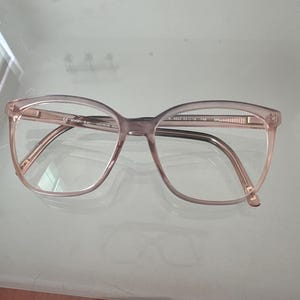 Può includere: Occhiali da vista rosa chiaro traslucido con una montatura di forma rettangolare arrotondata. Le aste interne presentano scritte stampate, tra cui il marchio JOOP! e le misure tecniche 53-18-140, indicanti una larghezza lente di 53mm e ponte di 18mm.