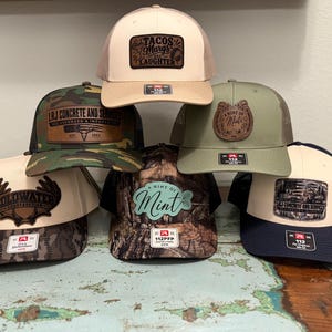 May include: A collection of six trucker hats featuring various designs, including camouflage, solid tan, olive green, and navy blue patterns. Each hat displays a unique leather or synthetic patch with text such as Tacos Margs and Laughter, LRJ Concrete and Services, Coldwater Construction, and A Hint of Mint.