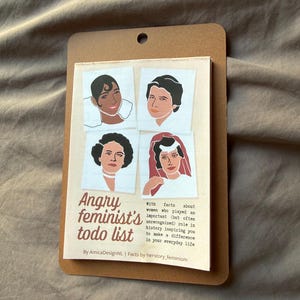 May include: A brown notepad with a printed "Angry feminist's todo list" and illustrations of four women. The text includes facts about women who played an important role in history. The notepad is on a brown fabric surface.