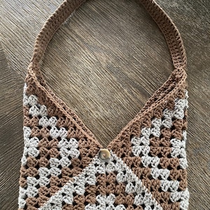 Peut inclure: Un sac à main en crochet avec un motif de carrés granny marron et gris clair. Le sac possède une anse marron unique et une ouverture supérieure en forme de V fermée par un bouton marron central. La structure est faite de mailles entrelacées.
