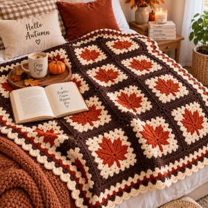 Könnte beinhalten: Eine handgefertigte Häkeldecke mit einem wiederkehrenden Muster aus quadratischen Motiven, die jeweils ein dunkelorangefarbenes Ahornblatt auf cremefarbenem Grund zeigen, umrahmt von dunkelbraunen Garnrändern mit gewelltem Abschluss.
